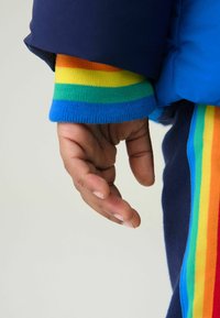 Kék puff dzseki mandzsettája szivárványcsíkos bordás kötésű részletekkel narancssárgában, zöldben, sárgában és kékben. Feltárt kéz nyúlik ki.