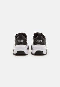 Zapatillas deportivas en blanco y negro con una parte superior texturizada, suela acolchada y un logotipo prominente en el talón trasero. Incluye una pestaña para tirar.