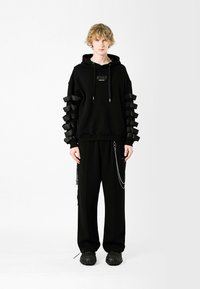 Sudadera con capucha negra y acentos texturizados en los hombros, con mangas largas y correas en capas. Combinada con pantalones negros y calzado grueso.