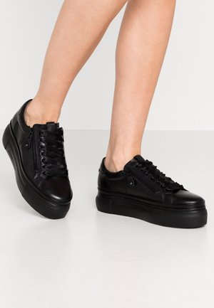 Sorte platformsneakers med sidelommer og satinbånd, båret på bare ben, stående på en hvid overflade og baggrund.