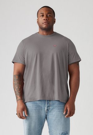 Camiseta de algodón gris con cuello redondo, mangas cortas y un pequeño logotipo rojo en el pecho. Corte holgado y textura suave. Combinada con jeans de color azul claro.
