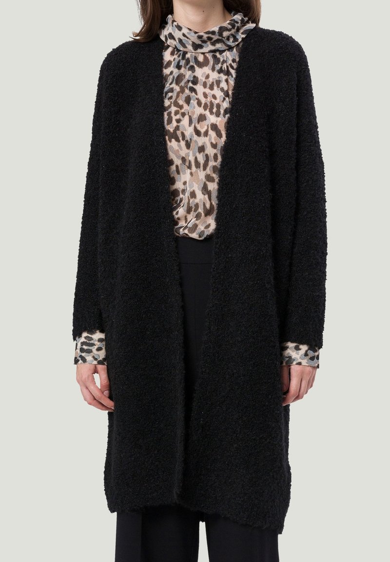 Femme portant un long cardigan noir texturé ouvert sur un chemisier à col haut imprimé léopard et un pantalon noir.