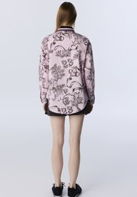 Różowa koszula z długim rękawem z brązowymi nadrukami botanicznymi i numerami. Posiada zaokrąglony dół, rękawy zapinane na guziki oraz klasyczny krój kołnierzyka.