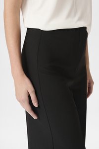 Černé kalhoty s hladkým, matným povrchem a přiléhavým střihem. Ruka spočívá na bočním švu, což ukazuje na minimalistický design.