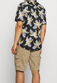 Zwarte shirt met korte mouwen met een tropisch wit en geel bloemenpatroon, gecombineerd met beige cargo shorts met zijzakken en een losse pasvorm.