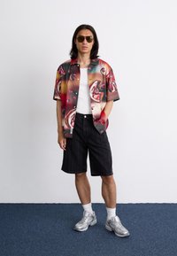 Korte mouwen overhemd met een kleurrijk abstract patroon, gecombineerd met zwarte pinstriped shorts en zilveren sneakers. Zonnebril op.