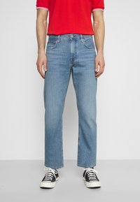 Jeans de mezclilla azul claro con un corte recto, cinco bolsillos y detalles de costura sutiles. Llevados con zapatillas negras y blancas.