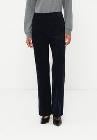 Tengerészkék kordbársony nadrág magas derékkal, egyenes szabású lábakkal és diszkrét varrási részletekkel. Fekete hegyes orrú cipővel párosítva.