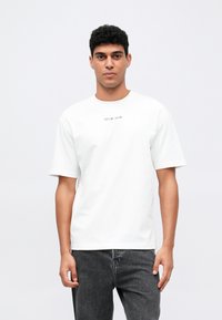 Hombre con cabello corto y rizado, vistiendo una camiseta blanca con un pequeño texto negro en el pecho y jeans gris oscuro, de pie frente a un fondo simple.