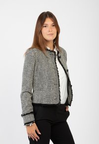 Giacca in tweed a spina di pesce grigia con bordi sfrangiati, due bottoni frontali e un design aderente. Abbinata a una camicia bianca e pantaloni neri.