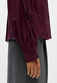 Top burdeos de manga larga con puños fruncidos, que presenta lazos laterales y un puño abotonado, combinado con pantalones de tela grises. Textura suave.