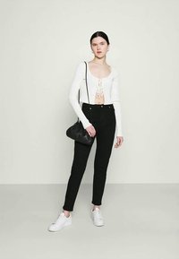 Jovem mulher com um top branco de manga longa cropped, calças pretas de cintura alta, ténis brancos, a segurar uma mala de ombro preta, contra um fundo liso.