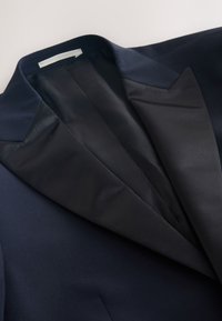 Blazer bleu marine avec un revers en satin à pointe et une doublure intérieure lisse. Épaules structurées et coupe ajustée qui renforcent le design formel.