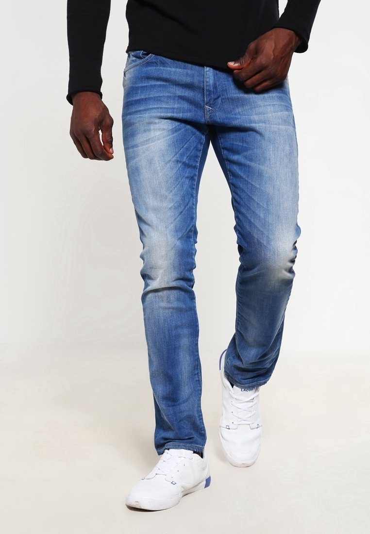 Vaqueros de mezclilla azul claro con un corte ajustado y un ligero desgaste. Tienen cinco bolsillos, cierre de cremallera y detalles de desgaste en las rodillas. Combinados con zapatillas blancas.