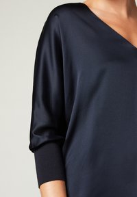 Blouse bleu marine avec un col en V, en tissu lisse. Présente des manches larges et des poignets ajustés. Texture douce, tombé raffiné.