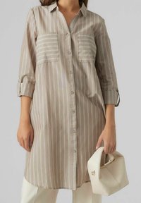 Woman in beige long-sleeve striped shirt dress holding a white soft handbag, wearing white pants against a plain background.