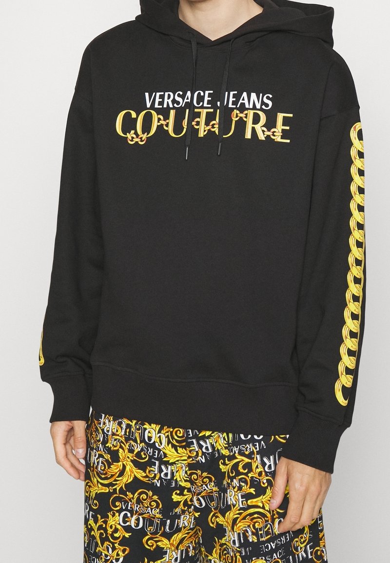 Sweat à capuche noir avec le texte « Versace Jeans Couture » et un motif chaîne dorée sur les manches, associé à un pantalon noir et jaune à motif.