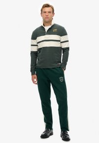 Grünes Langarm-Polo mit weißen Streifen und Logo, kombiniert mit grünen Jogginghosen, die einen weißen Seitenstreifen und ein Logo an der Oberschenkel haben.