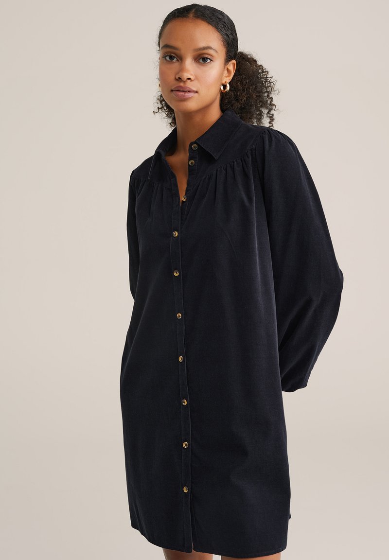 Rochie cambrată din corduroy bleumarin, cu guler, mâneci lungi puff și închidere cu butoane la fermoar. Are o croială lejeră și butoane de ton auriu.