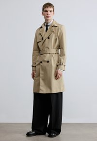 Béžový trenčkot z hladkej tkaniny, s dvojradovými gombíkmi, opaskom v páse a ramennými epolety. Štýlovo k širokým čiernym nohaviciam.