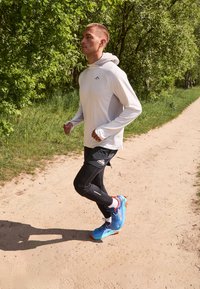 Felpa a maniche lunghe grigia chiara, leggings neri da corsa e scarpe sportive blu con dettagli arancioni, indossate da una persona che corre su un sentiero di terra.