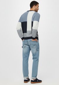 Man in een blauw, wit en zwart kleurgebroke sweater, lichte blauwe opgerolde jeans en zwarte sneakers, staande met zijn handen in zijn achterzakken.