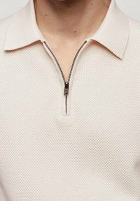 Polo en beige texturé avec une fermeture éclair à mi-hauteur, col ouvert, porté par une personne montrant le cou et le haut de la poitrine.