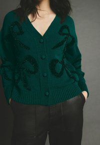 Tealfarbener Cardigan mit V-Ausschnitt, drei Knöpfen und strukturiertem Stoff mit schwarzen, glitzernden Rippmustern. Gerippte B�ündchen und Saum. Über schwarzen Lederhosen getragen.