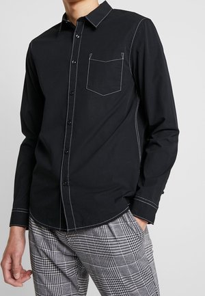 Mann trägt ein schwarzes Hemd mit weißen Kontrastnähten und graue karierte Hose, steht mit einer Hand in der Tasche.