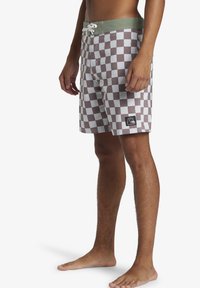 Brown and white checkered swim shorts with a green waistband, featuring a drawstring and a logo patch on the lower left side.