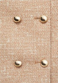 Tissu texturé couleur pêche avec un motif tissé, présentant trois boutons dorés brillants à surface lisse et un petit détail de fil près des boutons.