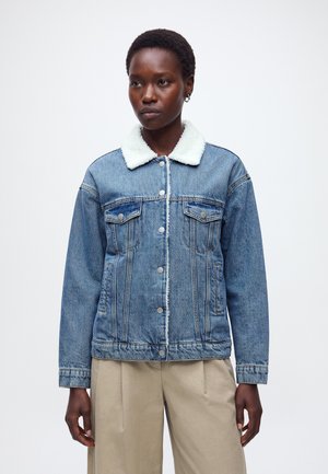 Femme portant une veste en jean bleue avec un col en polaire blanche et un pantalon beige à jambes larges, debout devant un fond blanc uni.