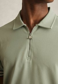 Tricou polo verde deschis cu închidere cu fermoar, material texturat și guler plat. Prezintă dungi orizontale pe mâneci.