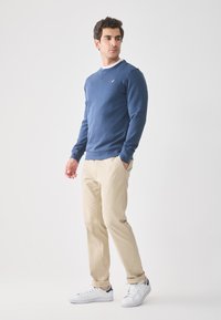 Mann som står og ser sideveis, iført en blå hettegenser, beige bukser og hvite sneakers mot en ensfarget hvit bakgrunn.