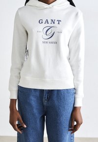 Bílá mikina s kapucí z bavlny, zdobená modrým logem s nápisy "GANT" a "NEW HAVEN." Slačuje se se světle modrými džíny, volného střihu.