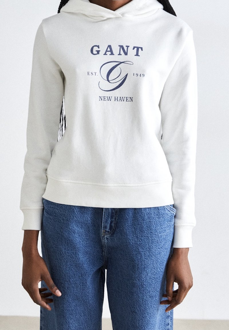 Bílá mikina s kapucí z bavlny, zdobená modrým logem s nápisy "GANT" a "NEW HAVEN." Slačuje se se světle modrými džíny, volného střihu.