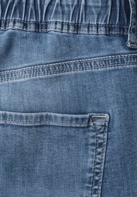 Back pocket detail of blue denim jeans with visible stitching and an elastic waistband section at the top.