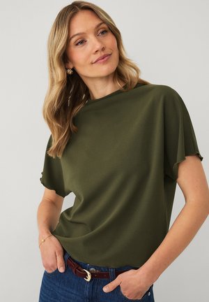 Kvinde med blond hår iført en olivengrøn kortærmet top, blå jeans, brun bælte og guldringeøreringe, står med hænderne i lommerne.