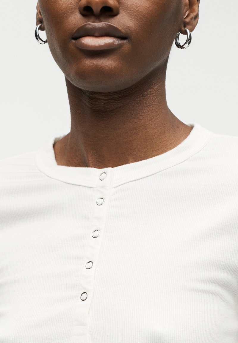 Gros plan d'une personne portant une chemise blanche boutonnée avec des boucles d'oreilles argentées en forme d'anneau, montrant le cou et le bas du visage.