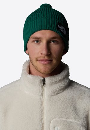 Gorro de punto verde con textura de canalé y un pompón en la parte superior, combinado con una chaqueta de forro polar blanca con cremallera y detalle de bolsillo.