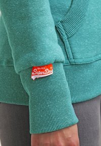 Sudadera color azul verdoso con puños acanalados, que presenta una etiqueta "SuperDry" en rojo y blanco. La textura del tejido es suave y con un patrón jaspeado.