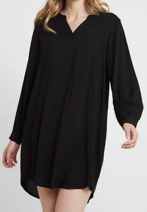 Femme portant une tunique noire ample à manches longues avec un col en V et une patte de boutonnage, debout devant un fond uni.