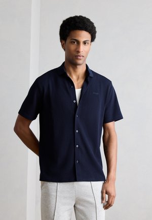 Άνδρας με κοντά σγουρά μαλλιά, φορώντας μπλε σκούρο πουκάμισο με κοντά μανίκια και ανοιχτό γκρι παντελόνι, στέκεται μπροστά σε απλό φωτεινό φόντο.