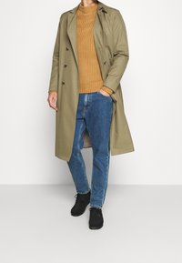 Trench-coat olive porté sur un pull moutarde côtelé, associé à un jean bleu et des chaussures noires. Présente un devant boutonné et des poches latérales.
