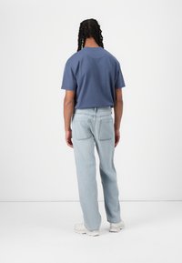 Avslappet blå T-skjorte med korte ermer, kombinert med lyseblå rettkuttjeans og hvite joggesko, fotografert fra baksiden.