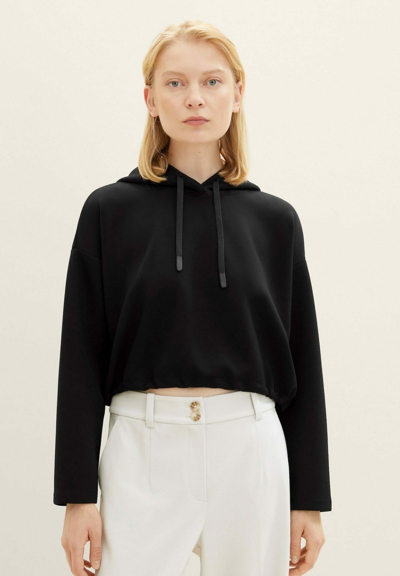 Schwarze, cropped Kapuzenjacke mit Kordelzug, langen Ärmeln und dezenter Struktur. Kombiniert mit weißen Hosen mit doppeltem Knopfverschluss.