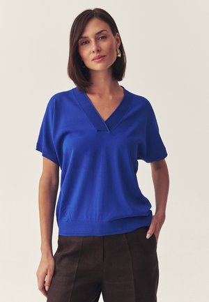 Woman with short dark hair wearing gold earrings, a blue short-sleeve V-neck top, and dark brown pants, posing with one hand in pocket.