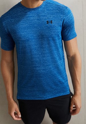 Man draagt een blauw atletisch shirt met korte mouwen met subtiele patronen en een zwart logo, gecombineerd met zwarte shorts, staand tegen een effen muur.