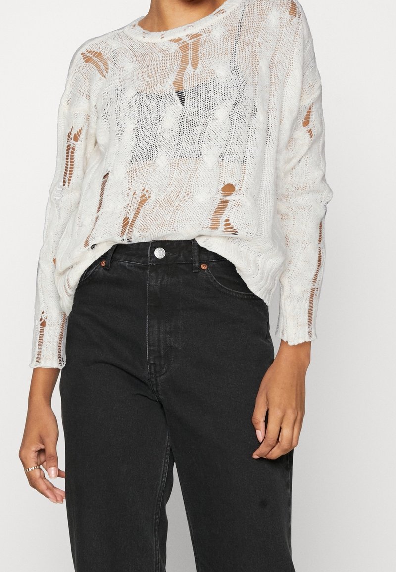Personne portant un pull en maille blanche usé sur un sous-vêtement noir, associé à un jean noir taille haute.