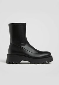 Botte en cuir noire mi-mollet avec semelle épaisse à crampons et bout arrondi, vue de profil sur fond uni.